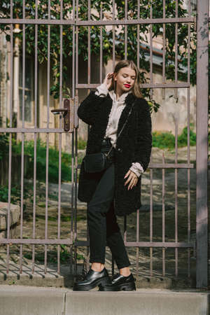 Close-up photo of young beautiful woman in black fur coat, jeanse and shoes posing on streetの写真素材