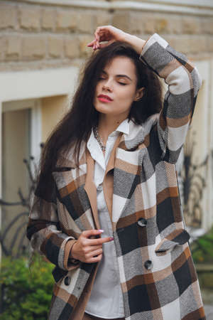 Closeup portrait of young beautiful fashionable woman wearing checkered long coat, beige pants and white blouse . Lady posing on city street.の写真素材