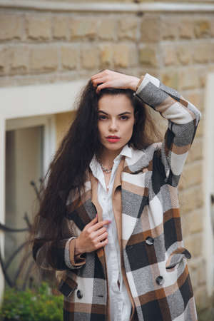 Closeup portrait of young beautiful fashionable woman wearing checkered long coat, beige pants and white blouse . Lady posing on city street.の写真素材