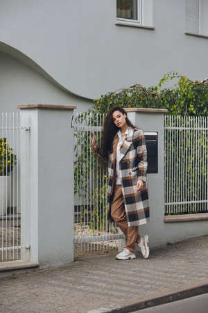 Closeup portrait of young beautiful fashionable woman wearing checkered long coat, beige pants and white blouse . Lady posing on city street.の写真素材