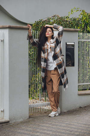 Closeup portrait of young beautiful fashionable woman wearing checkered long coat, beige pants and white blouse . Lady posing on city street.の写真素材