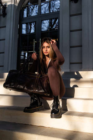 Fashionable young brunette woman with long legs wearing Solid Long Sleeve Bodycon One Piece Jumpsuits posing with a leather brown travel bagの写真素材