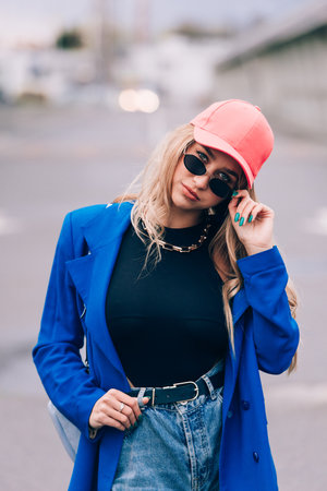 Young sexy blonde hipster woman posing on the street. Wearing blue stylish jacket, jeans and baseball hat and sunglasses.の写真素材