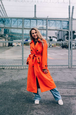 Fashionable blonde woman wearing orange coat, blue jeanse , white sneackers and neckchain posing in streetの写真素材