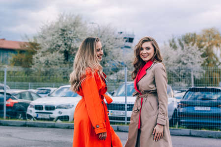 Fashionable happy smiling blonde woman wearing orange coat, blue jeanse and neckchain communicate with her girlfriend on the street.の写真素材