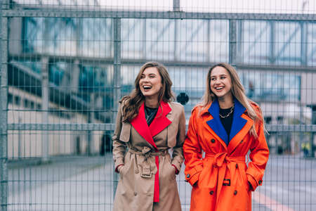 Fashionable happy smiling blonde woman wearing orange coat, blue jeanse and neckchain communicate with her girlfriend on the street.の写真素材