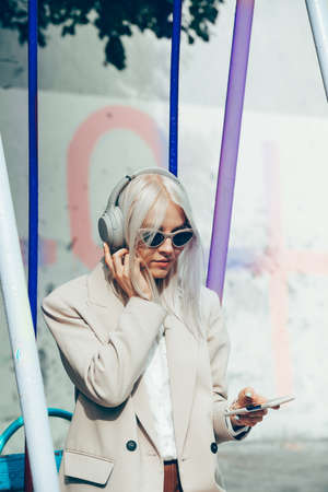 Portrait of blonde woman in headphones listening music with sunglasses. Hipster fashion model wear...