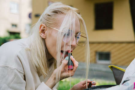 young smiling hipster happy woman doing make-up using lipstick and mirror outdoorsの写真素材