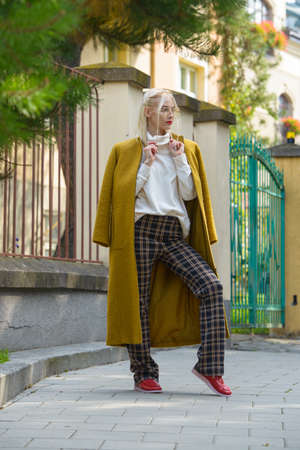 Fashionable beautiful young woman with blond hair in a stylish long coat, checkered pants, red shoes and glasses poses in the city streets.の写真素材
