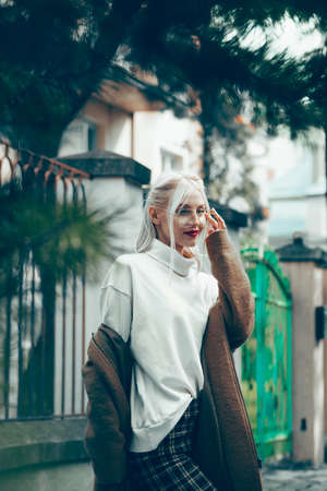 Fashionable beautiful young woman with blond hair in a stylish long coat, checkered pants and glasses poses in the city streets.の写真素材