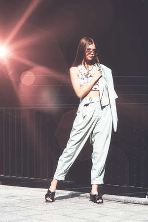Fashion portrait of young woman wearing sunglasses, top, slingbacks, blue suit. Young beautiful happy model posing near gray metal gridの写真素材