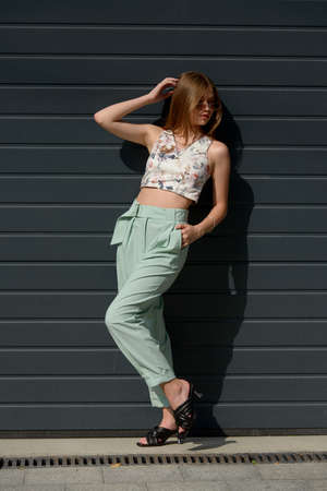 Fashion portrait of young woman wearing sunglasses, top, slingbacks, blue suit. Young beautiful happy model posing near gray metal gridの写真素材