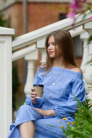 A female posing near flower on a stone curb while drinking coffe. Girl wearing a striped jumpsuitの写真素材