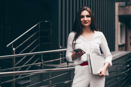 Beautiful young caucasian businesswoman in a white clothes with a laptop and notebook outdoor near business centreの写真素材