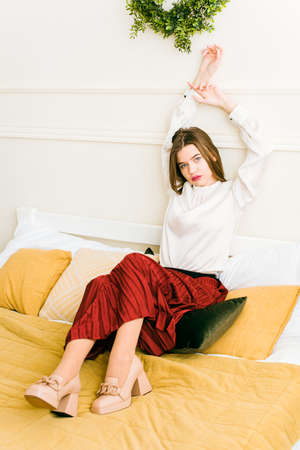 Portrait of fashionable women in red skirt, white blouse and stylish beige high-heeled shoes with a chain buckle posing in a bedの写真素材