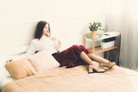 Portrait of fashionable women in red skirt, white blouse and stylish beige high-heeled shoes with a chain buckle posing in a bedの写真素材
