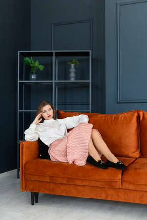 Portrait of fashionable young woman in a beige skirt, white blouse and stylish beige black seude shoes with a buckle posing on a orange sofa.の写真素材