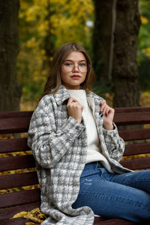 Close-up portrait of young woman in a stylish trench coat and in a white sweater resting on the bench. warm autumn day.の写真素材