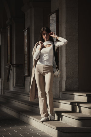 beautiful brunette girl dressed in white blouse, pants, sneakers with a handbag and camel coat in hands. Stylish trendy fashion outlookの写真素材