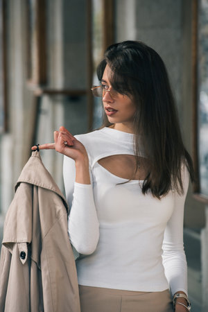 beautiful brunette girl in a sunglasses dressed in white blouse with a beige coat in hands. Stylish trendy fashion outlookの写真素材