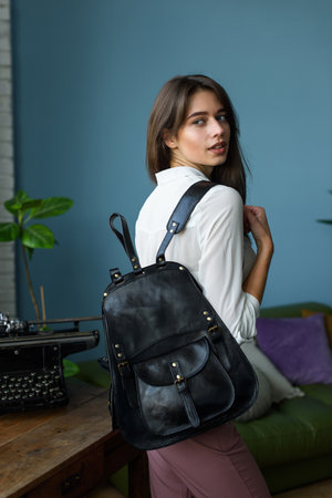 beautiful slender girl in burgundy pants and a white blouse posing with a black leather backpack