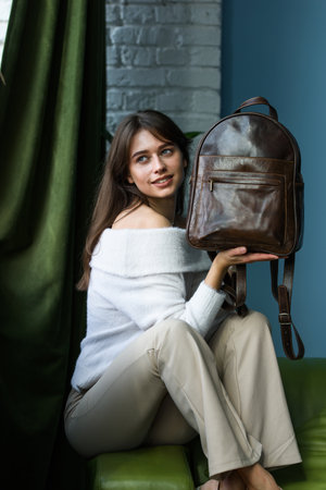 beautiful slender girl in beige pants and olive sweater posing while sitting on a green sofa with a small shiny brown leather backpackの写真素材