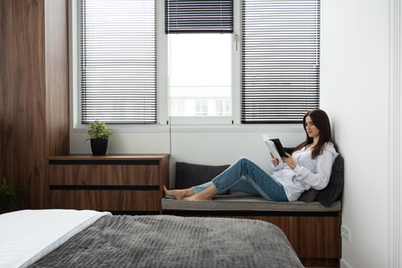 Woman reading book in a bedroom with bed and wooden wardrobe in modern apartmentの写真素材
