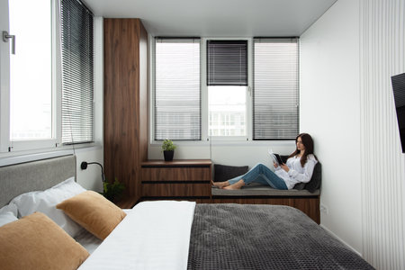 Woman reading book in a bedroom with bed and wooden wardrobe in modern apartmentの写真素材