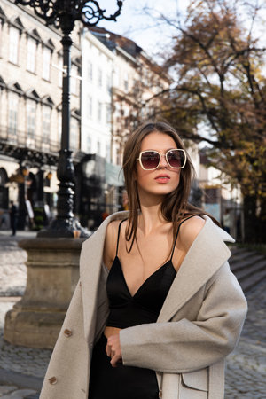charming brunette posing on the street in spring, wearing a stylish beige coat, a top with razors and sunglassesの写真素材
