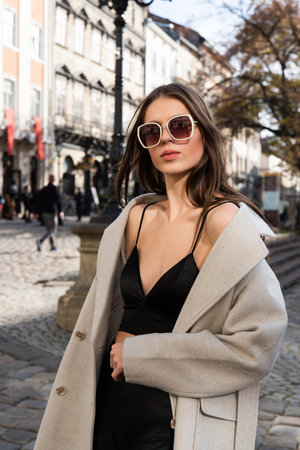 charming brunette posing on the street in spring, wearing a stylish beige coat, a top with razors and sunglassesの写真素材
