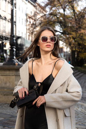 charming brunette posing on the street in spring, wearing a stylish beige coat, a top with razors and sunglassesの写真素材