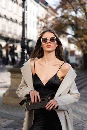 charming brunette posing on the street in spring, wearing a stylish beige coat, a top with razors and sunglassesの写真素材