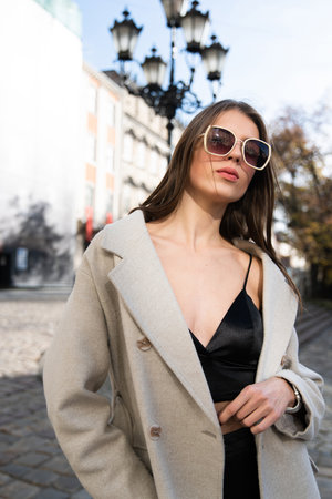 charming brunette posing on the street in spring, wearing a stylish beige coat, a top with razors and sunglassesの写真素材