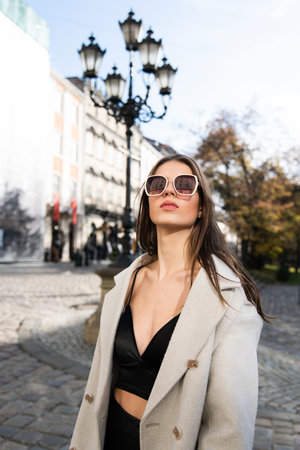 charming brunette posing on the street in spring, wearing a stylish beige coat, a top with razors and sunglassesの写真素材