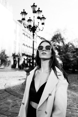 charming brunette posing on the street in spring, wearing a stylish beige coat, a top with razors and sunglassesの写真素材