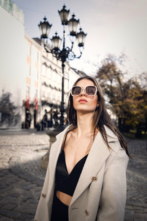 charming brunette posing on the street in spring, wearing a stylish beige coat, a top with razors and sunglassesの写真素材