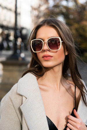 close-up portrait of a charming brunette on the street in spring, wearing a stylish beige coat, a top with razors and sunglassesの写真素材