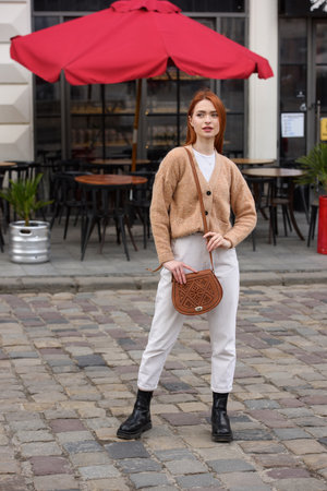 a red-haired girl in white pants and a beige sweater poses outside with a small leather handbagの写真素材