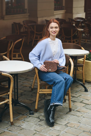 a red-haired girl in a blue jeans and a sweater poses outside with a small leather handbagの写真素材