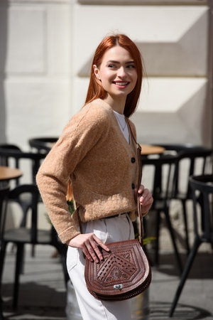 a red-haired girl in white pants and a beige sweater poses outside with a small leather handbagの写真素材