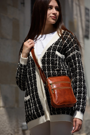 photo of beautiful woman in a white leggings, T-shirt and sweater posing with small brown leather bagの写真素材
