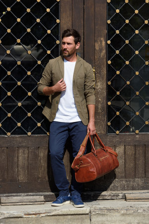 a man with a bag. black leather travel bag, street photo. man is wearing blue sneakers, jeans, a white T-shirt and a jacketの写真素材