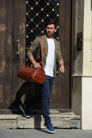 a man with a bag. black leather travel bag, street photo. man is wearing blue sneakers, jeans, a white T-shirt and a jacketの写真素材