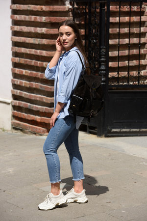 Beautiful young girl wearing blue jeans, shirt and white t-shirt posing with black leather backpackの写真素材
