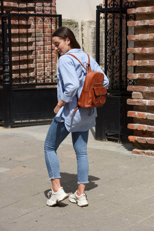 Beautiful young girl wearing blue jeans, shirt and white t-shirt posing with brown leather backpackの写真素材