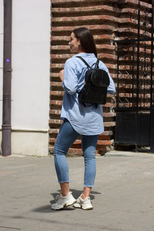 Beautiful young girl wearing blue jeans, shirt and white t-shirt posing with black leather backpackの写真素材