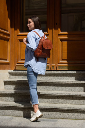 Beautiful young girl wearing blue jeans, shirt and white t-shirt posing with brown leather backpackの写真素材