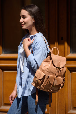Beautiful young girl wearing blue jeans, shirt and white t-shirt posing with beige leather backpackの写真素材