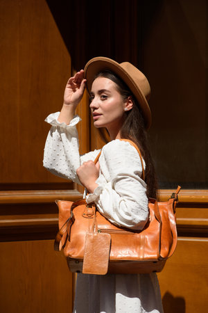 brunette with long hair wearing white romantic summer dress and beige hat posing with luxury leather bagの写真素材