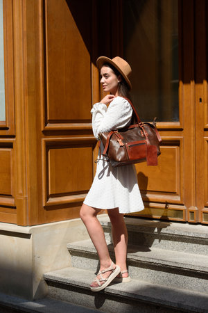 brunette with long hair wearing white romantic summer dress and beige hat posing with luxury leather bagの写真素材